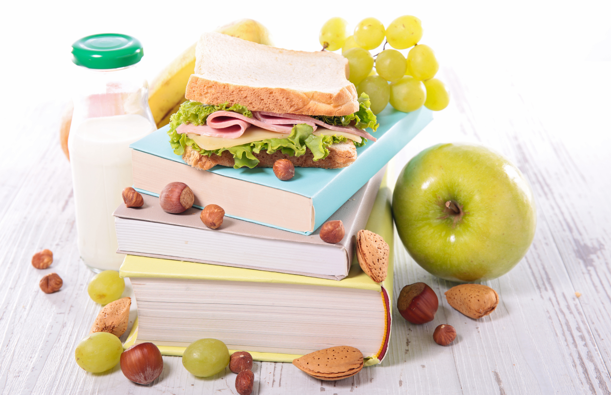 Stack of 3 books with a sandwich on top, apples, grapes, glass of milk, and nuts