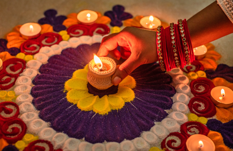 A mandala made of sand in different colors, on the ground, with a hand reaching down to place a lit candle in the center.