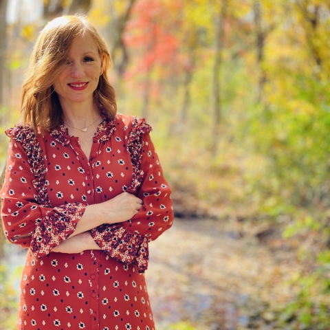 Red headed woman wearing a red dress with arms crossed and smiling in the forest with the leaves changing colors to red and yellow.